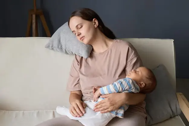 A grandparent gently holding a baby while the exhausted mother sleeps in a nearby chair