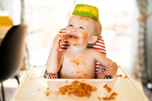 A happy, messy baby in a high chair, smiling with a variety of colorful, safe, mashed foods on the tray